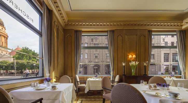 An opulent restaurant interior features multiple set tables amidst wood-paneled walls and gold trim. Large windows overlook a busy city street with buildings and trees.