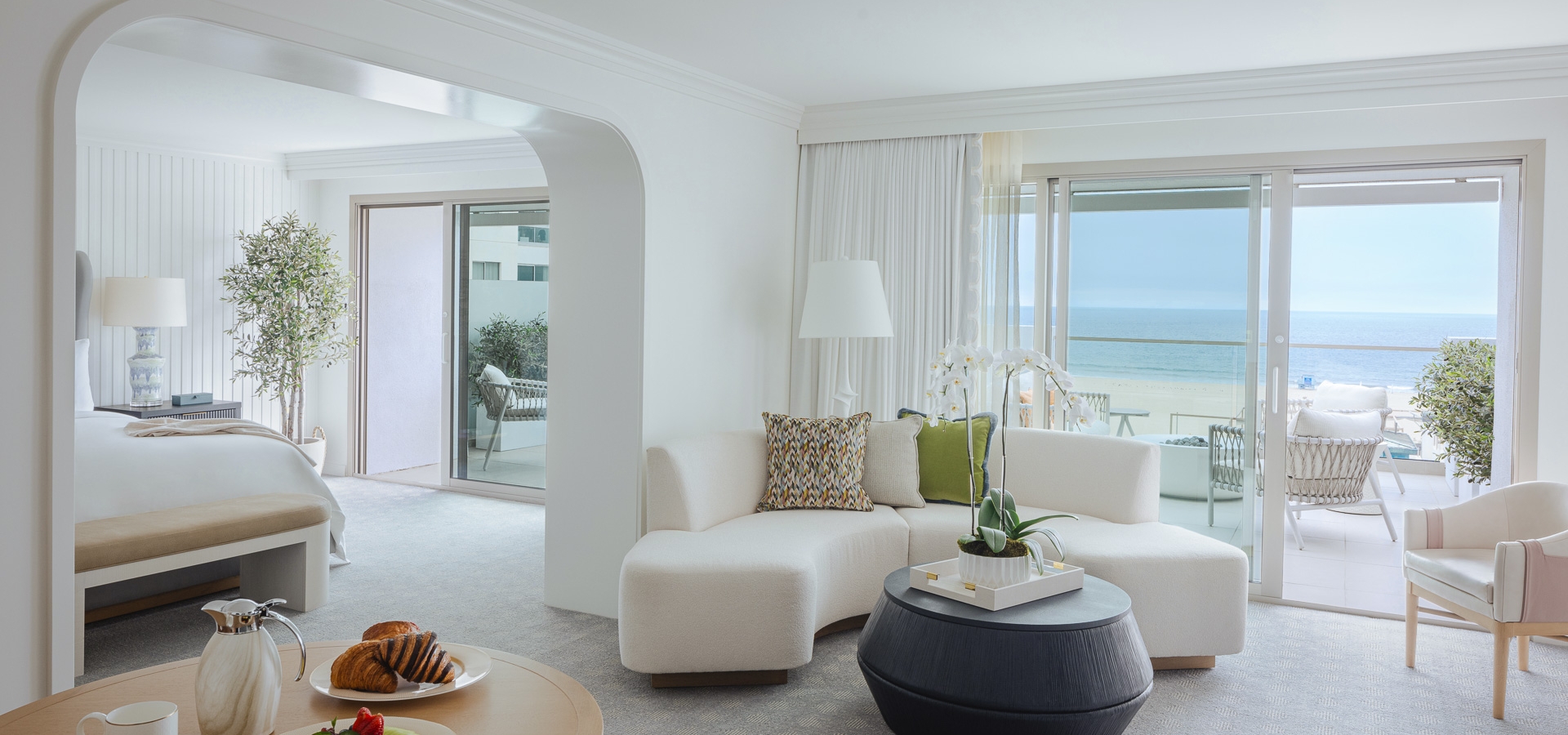 A breakfast spread of fruit and croissants sits on a wooden table in a bright, modern hotel suite, overlooking an ocean-view balcony.
