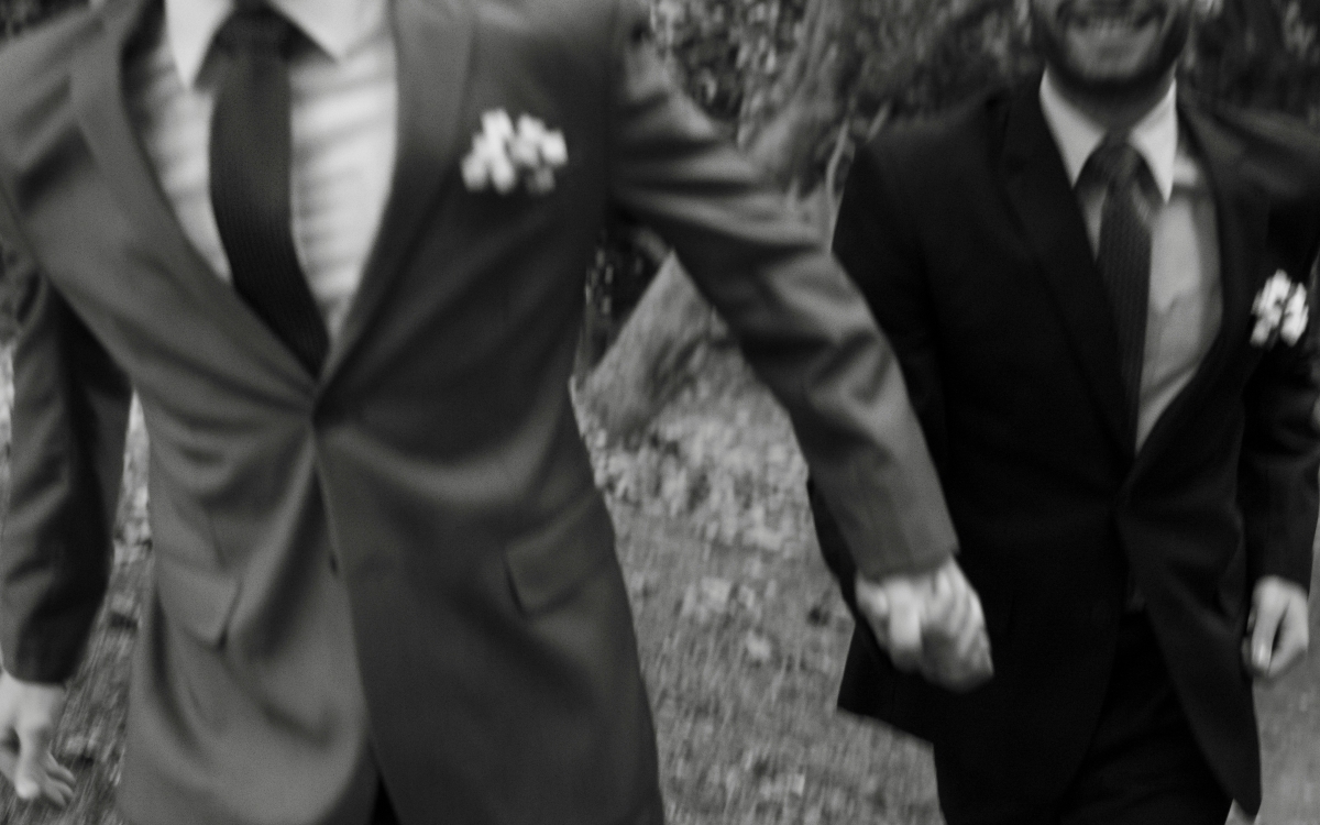 A close-up view of two people’s hands gently resting on top of each other. The hands wear gold and diamond wedding and engagement rings and a classic men’s silver watch, resting against the bride’s lace dress.