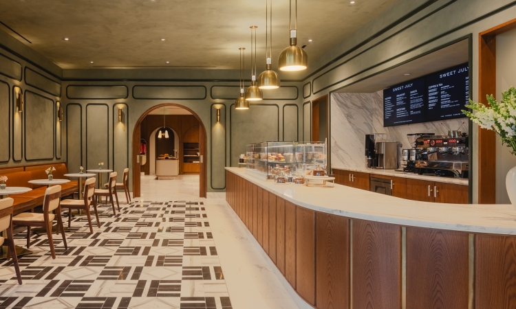 A modern café interior showcases a long, curved pastry counter and espresso bar, flanked by a dining area with sage green paneled walls and patterned floor.