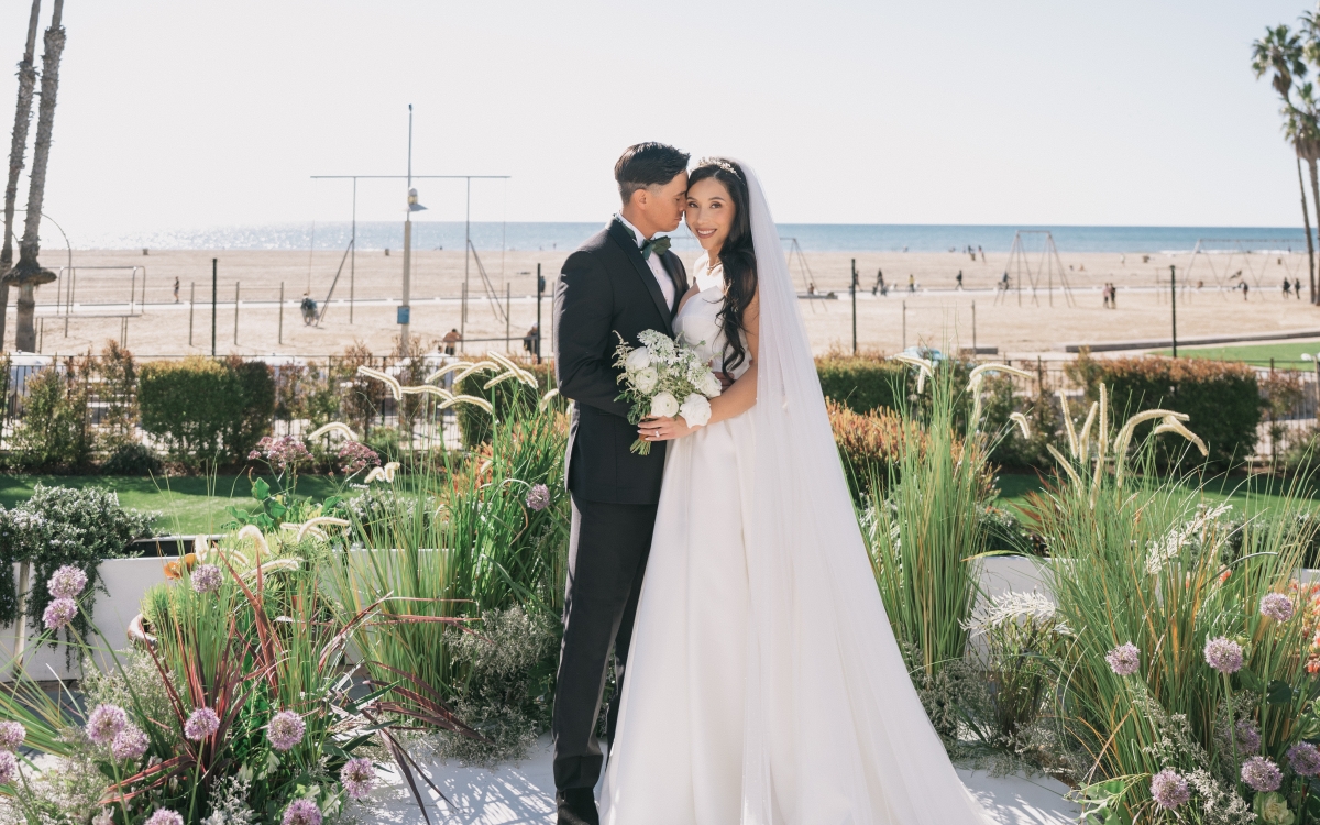 A bride and groom stand and embrace on a white circular stage outdoors on a sunny day. They are framed by tall wild purple flowers and grasses, with a beach, ocean, and palm trees in the background.