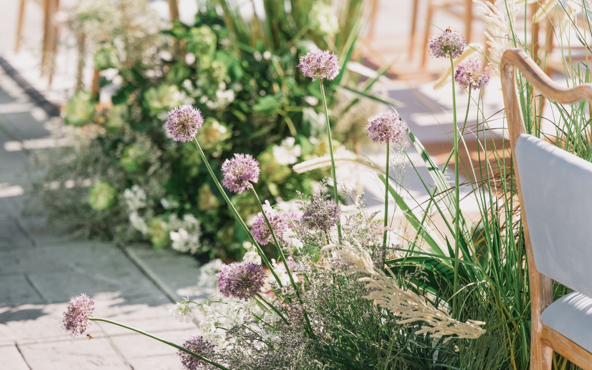 A detailed close-up of a wedding aisle runner, featuring a series of low-level, natural garden-style floral arrangements with purple allium and feathery grasses. Paved stone and wooden chairs are visible to the sides.