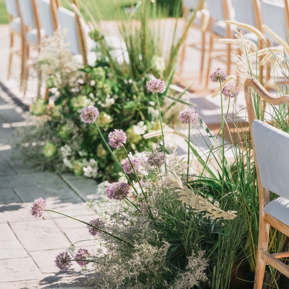 A detailed close-up of a wedding aisle runner, featuring a series of low-level, natural garden-style floral arrangements with purple allium and feathery grasses. Paved stone and wooden chairs are visible to the sides.