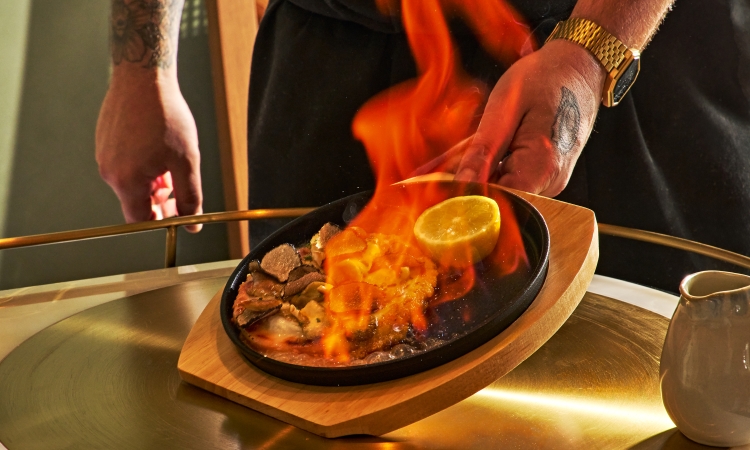 A cast-iron dish with food and a lemon half is flambéed, vibrant flames rising high. A person’s hands are visible, serving it on a wooden board atop a golden cart.