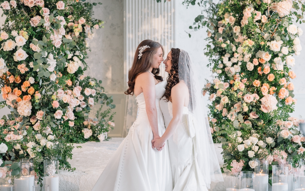 Two women in white wedding dresses, stand holding hands and smiling under a large, lush floral arch made of roses and eucalyptus. They are surrounded by glowing candles in glass holders on the floor.