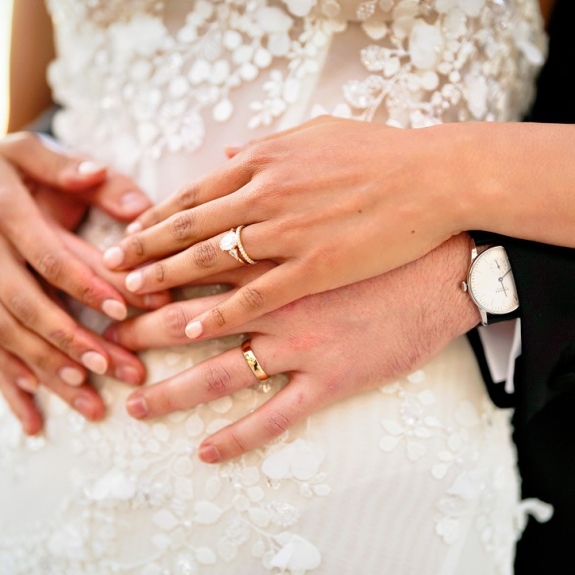 A close-up view of two people’s hands gently resting on top of each other. The hands wear gold and diamond wedding and engagement rings and a classic men’s silver watch, resting against the bride’s lace dress.