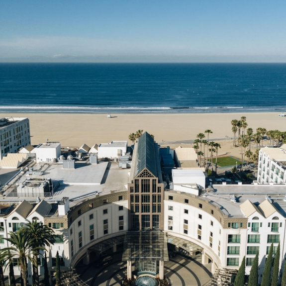 A large building complex, featuring a central structure with a glass roof, overlooks a wide sandy beach meeting the vast blue ocean, surrounded by other urban buildings and palm trees.