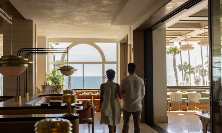 A couple walks into a luxurious restaurant, featuring warm brass lights, leather seating, and large arched windows overlooking the ocean and palm trees.